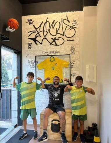 O Tio C sentado entre dois alunos no estúdio, com a camisa da seleção na parede