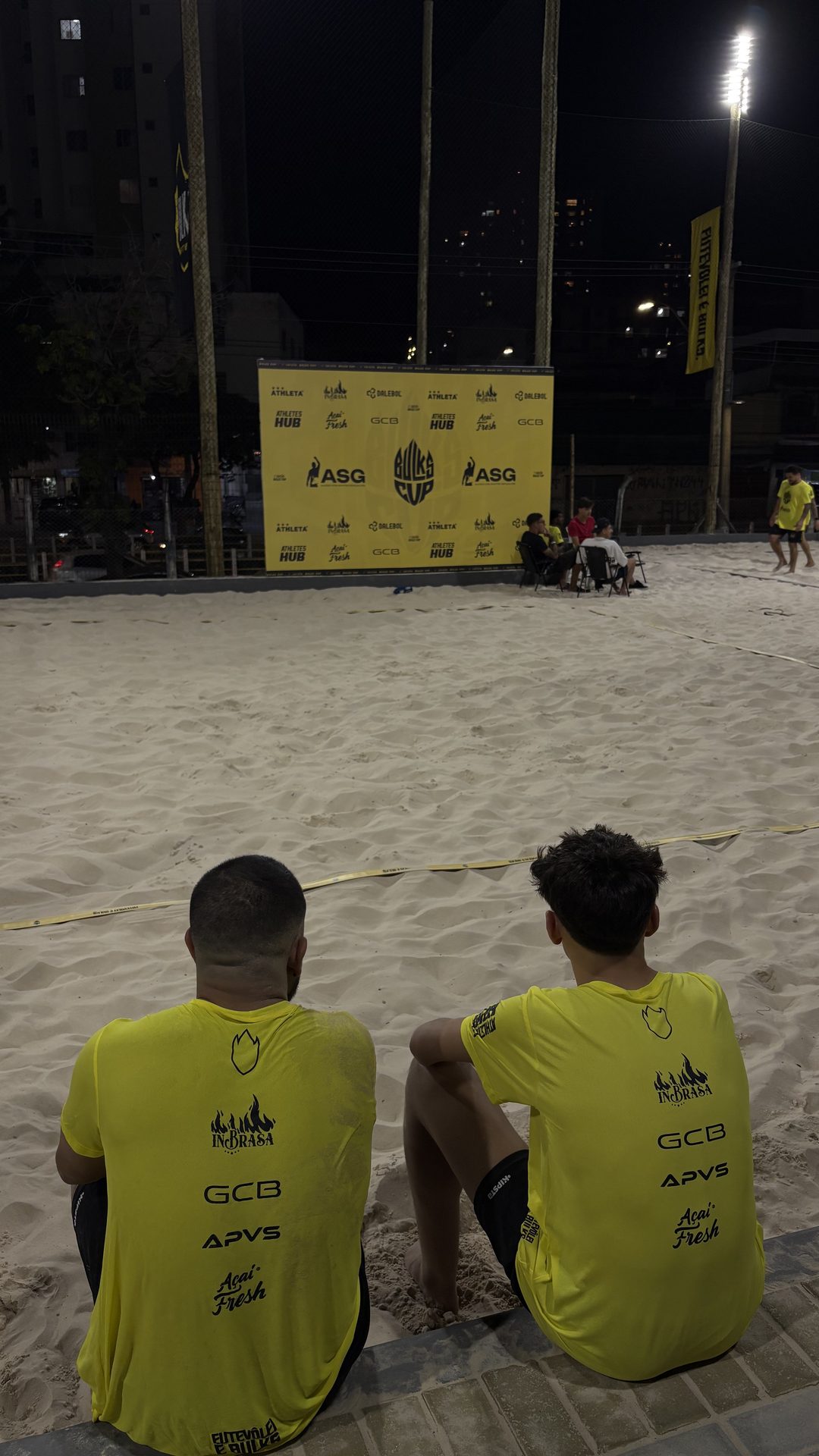 Dois iniciantes sentados na beira da quadra, à noite, olhando o jogo de fora